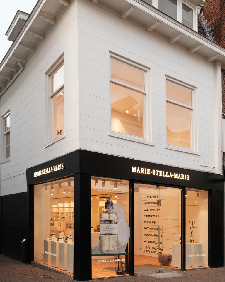 Exterior view of a Marie-Stella-Maris store located on a street corner. The building has a clean, modern design with white upper walls and black accents around the ground-floor storefront. Large glass windows display neatly arranged products and a large bottle graphic inside. The illuminated “MARIE-STELLA-MARIS” signage appears above the entrance and windows, and the warm interior lighting creates an inviting atmosphere.