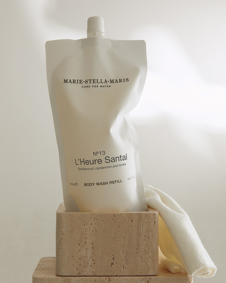 Marie-Stella-Maris No.13 L'Heure Santal Body Wash Refill (500 ml) in a minimalist white refill pouch displayed on a natural stone pedestal with a soft towel. The refill packaging highlights the signature fragrance with notes of sandalwood, cardamom and tonka.