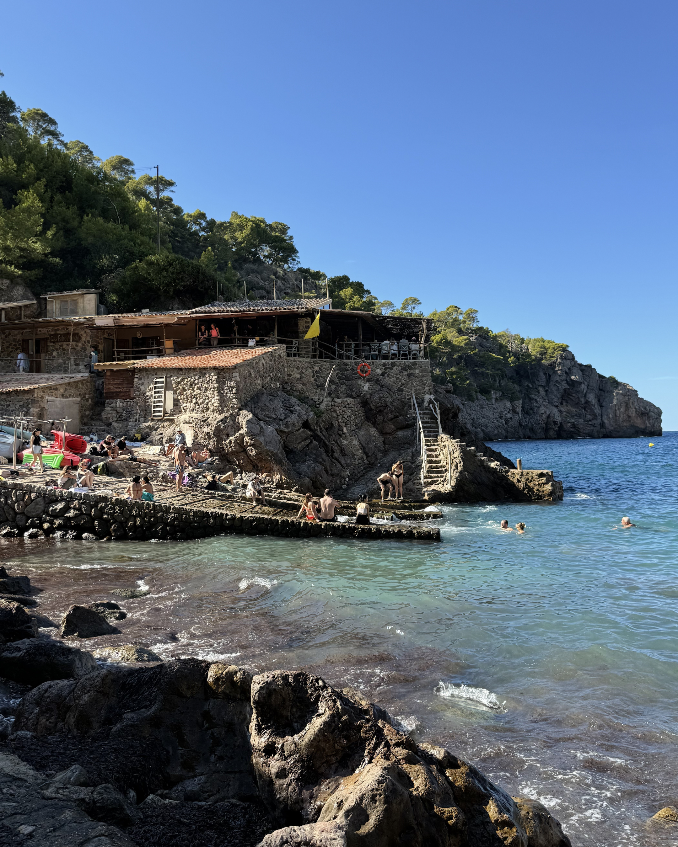 Visitors swimming and relaxing along the rocky shoreline of Cala Deià, Mallorca, with stone buildings and clear blue Mediterranean waters.