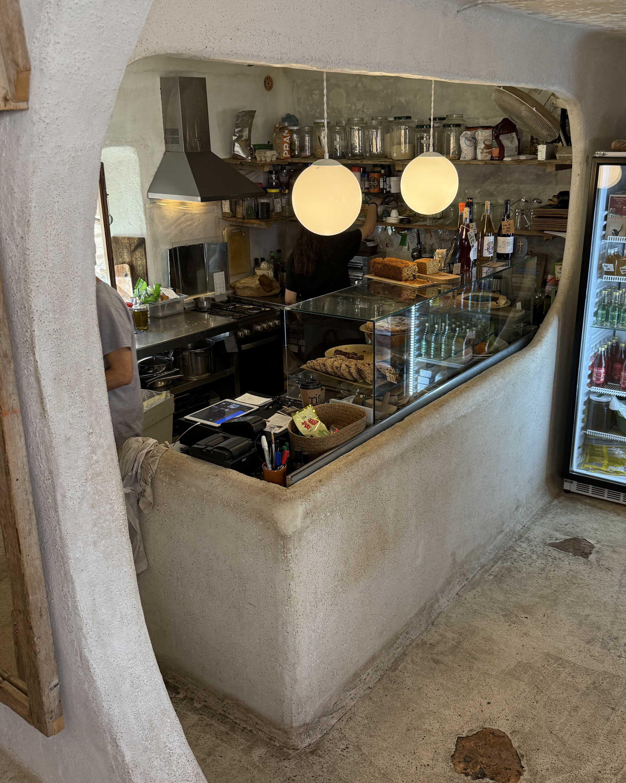 Interior of De Monio café in Mallorca, featuring a rustic open kitchen, homemade cakes in a glass display and warm pendant lighting.