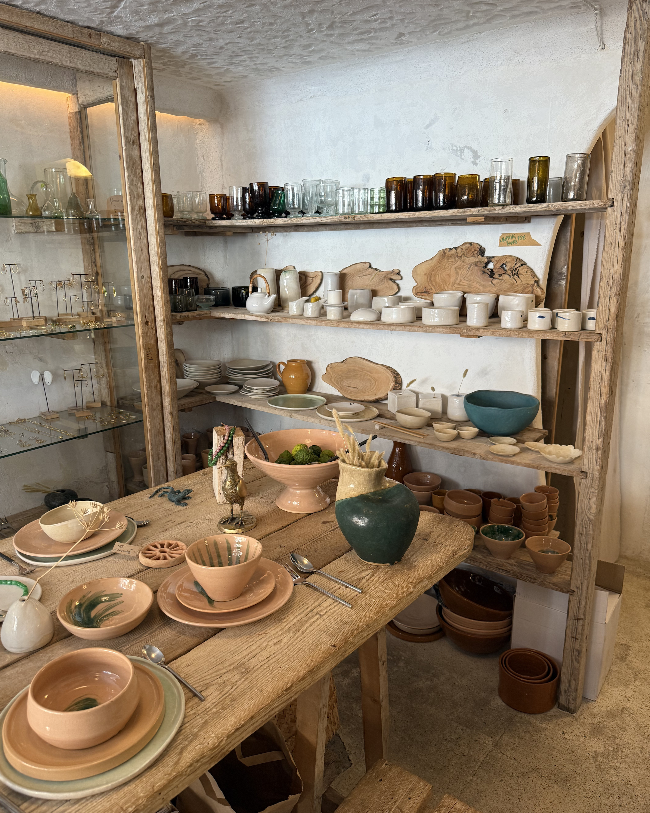 Interior of De Monio in Mallorca featuring handcrafted ceramics, wooden serving boards and colourful glassware on rustic wooden shelves.