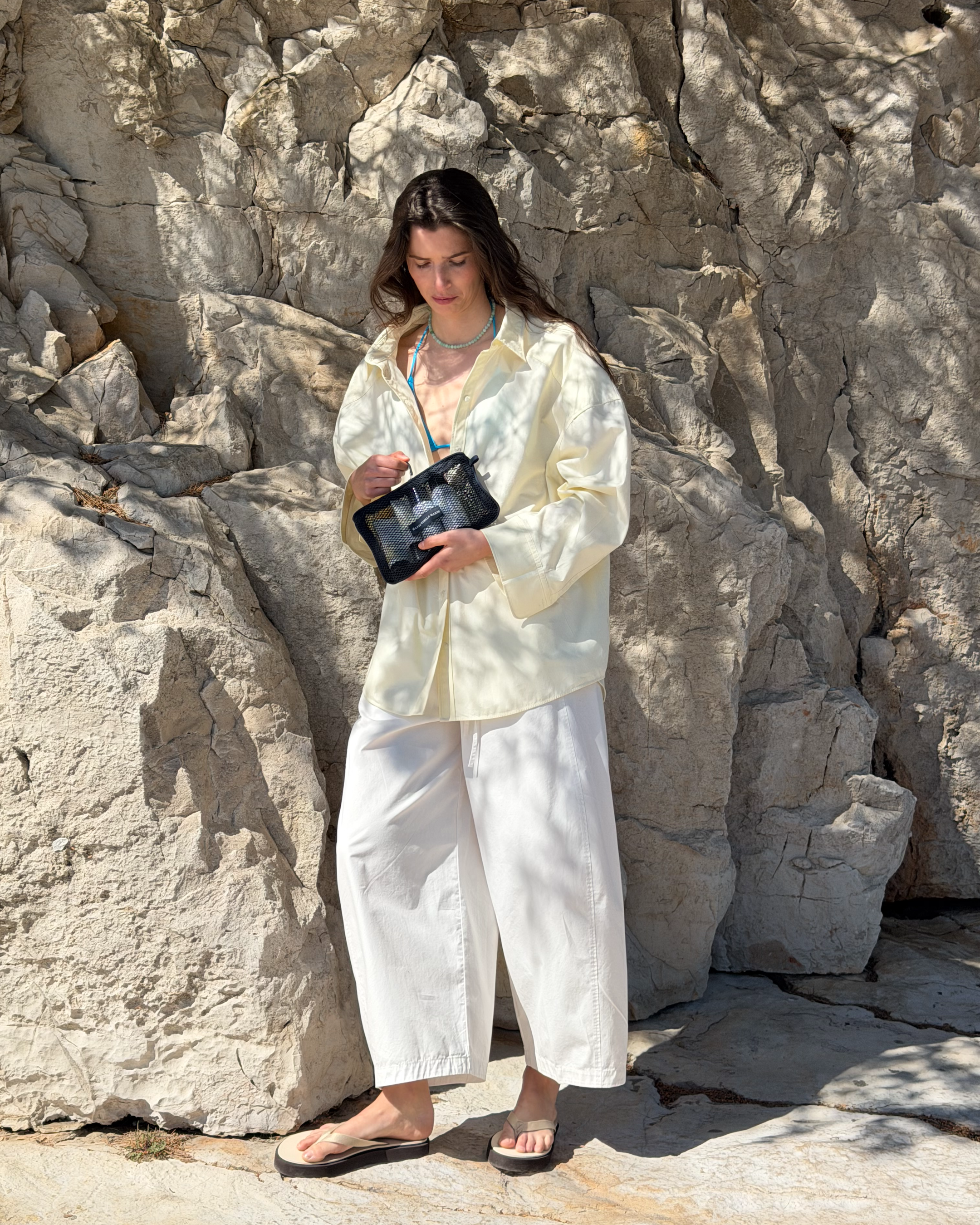 Person standing beside the limestone cliffs of Cala Deià, holding a Marie-Stella-Maris mesh pouch and dressed in relaxed summer layers.