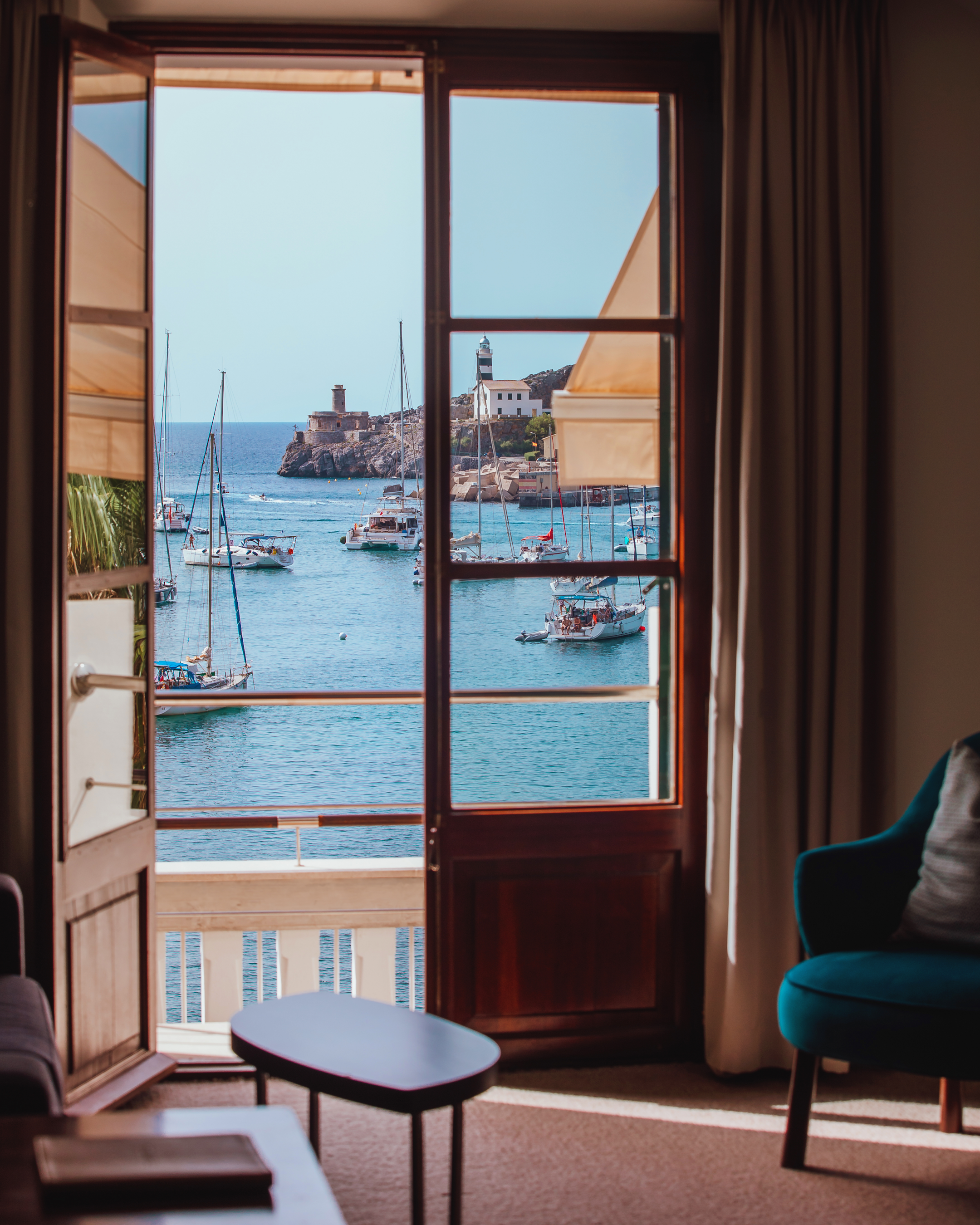 Open balcony doors overlooking Port de Sóller harbour in Mallorca, with sailboats, a lighthouse and turquoise sea on a sunny day.