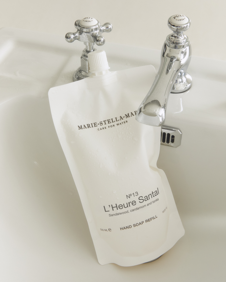 Marie-Stella-Maris No.13 L'Heure Santal Hand Soap Refill (500 ml) in a minimalist white refill pouch displayed in a sink beneath a classic chrome tap. Water droplets on the refill pouch highlight the refillable hand care routine, featuring the signature fragrance with notes of sandalwood, cardamom and tonka.