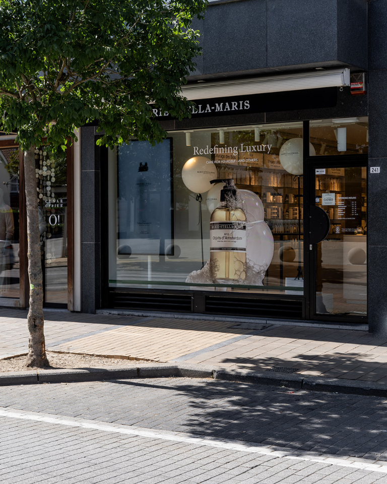 Exterior of a Marie-Stella-Maris store with a large display window showcasing an oversized product bottle installation and branded balloons. The storefront features black signage, glass doors, and the text ‘Redefining Luxury’ on the window, with a tree-lined sidewalk in front.