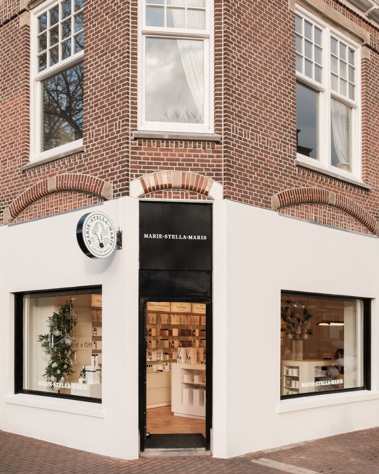 Corner storefront of the Marie-Stella-Maris boutique in Laren—white-painted facade on a brick building, large display windows, a black entrance door, and the Marie-Stella-Maris logo sign above the entrance, with the warmly lit interior visible through the windows.
