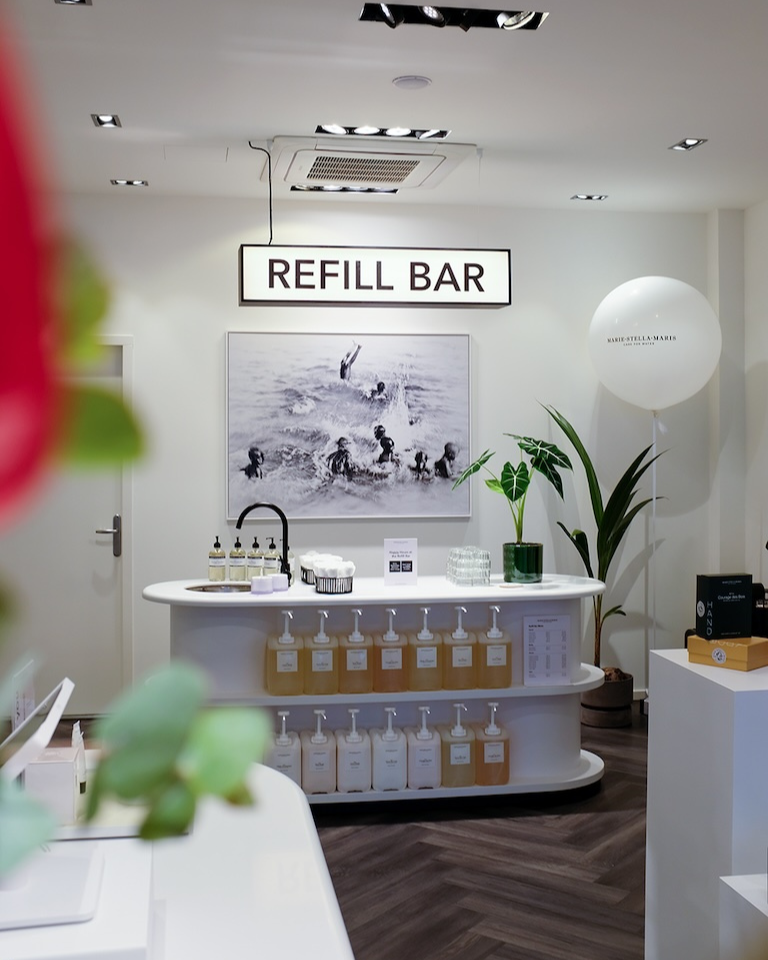 Interior of a Marie-Stella-Maris store featuring a white refill bar with multiple refill bottles, a sink with pump dispensers, and a sign reading ‘REFILL BAR’ above it. A black-and-white ocean photograph hangs on the wall behind the counter, with plants and a branded white balloon adding a clean, modern atmosphere.
