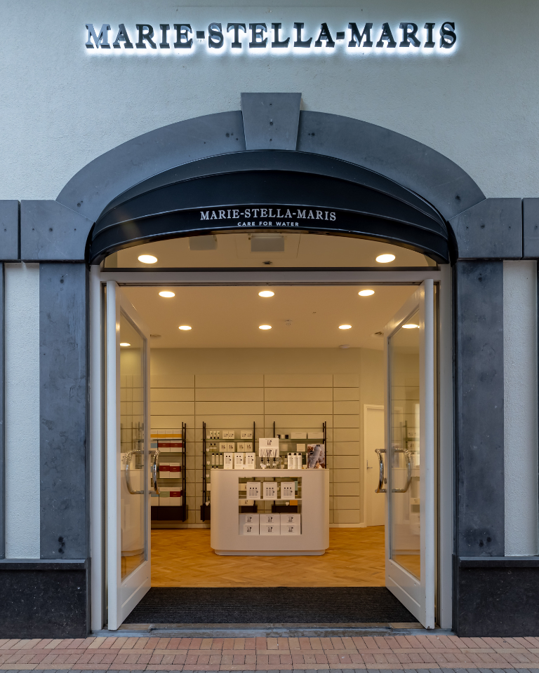Inside the Marie-Stella-Maris boutique at Designer Outlet Roermond: large ‘MARIE-STELLA-MARIS – Care for Water’ wall sign above black-framed glass shelves of home fragrance and body care, with a curved white display island on herringbone flooring.