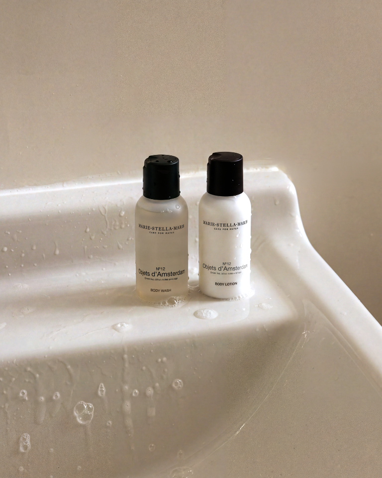 Travel-size Marie-Stella-Maris No. 12 Objets d’Amsterdam body wash and body lotion standing on a wet shower ledge in warm sunlight.