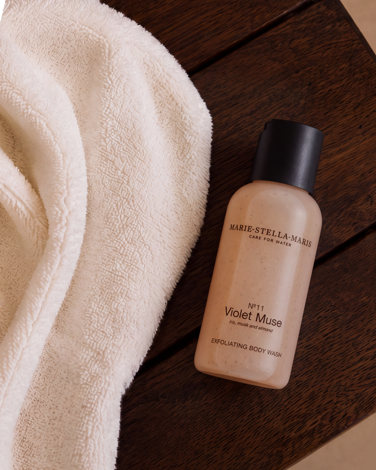 Travel-size Marie-Stella-Maris No. 11 Violet Muse exfoliating body wash displayed on a dark wooden bench beside a soft white towel.