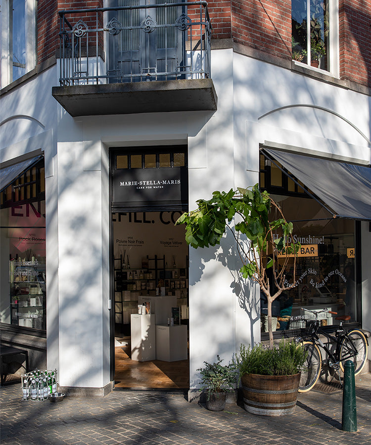 Corner storefront of the Marie-Stella-Maris boutique in Breda—white facade beneath an iron balcony, window awnings, planters and a bicycle outside, and ‘Refill Bar’ signage visible in the window.