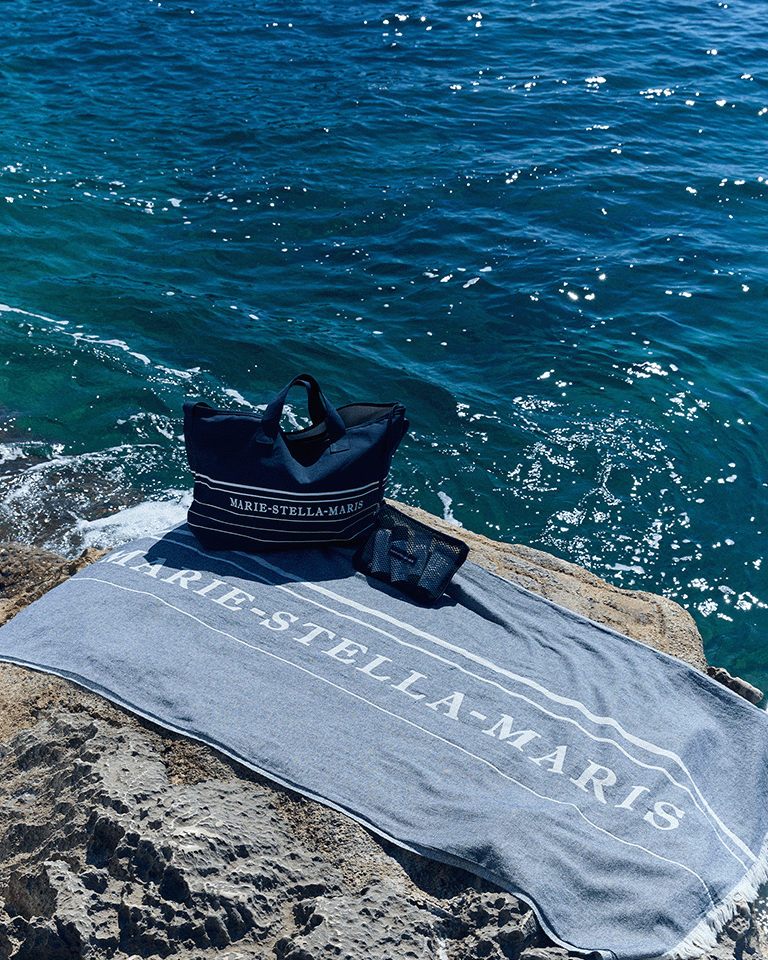 Marie-Stella-Maris beach setup with a towel laid on a rocky shore, paired with the limited edition summer 2026 navy blue tote bag and black mesh pouch—part of the limited edition gift with purchase collection—overlooking clear sparkling sea water.
