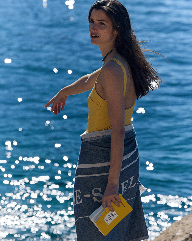 Person standing by the sea wearing a yellow swimsuit and wrapped in a Marie-Stella-Maris towel, holding a yellow book, with sunlight reflecting on the water in the background.