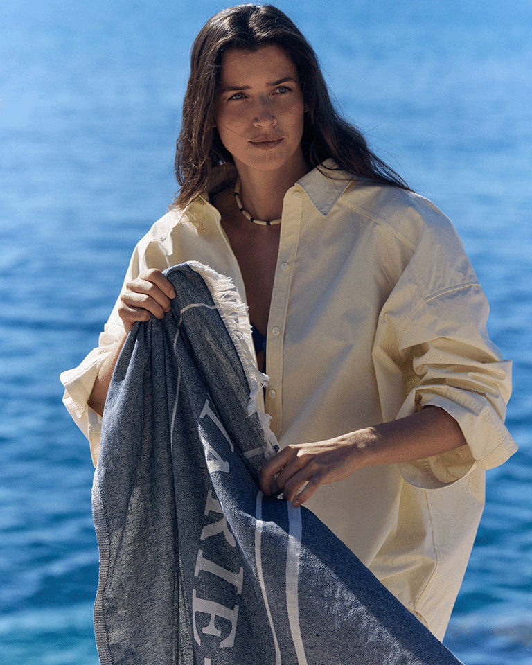 Person standing by the sea wearing a light shirt and holding a Marie-Stella-Maris towel, part of the limited edition gift with purchase collection, with sparkling blue water in the background.