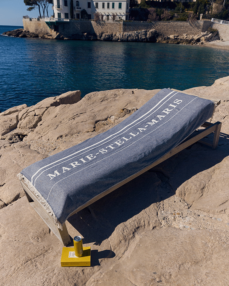 Marie-Stella-Maris towel draped over a sunbed on a rocky shoreline, part of the limited edition gift with purchase collection, with a yellow book and product placed nearby and the sea and coastal buildings in the background.