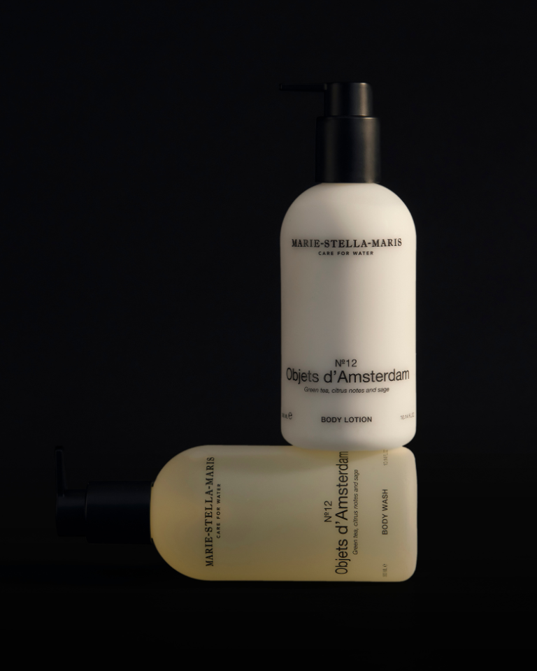 Marie-Stella-Maris Body Lotion No.12 Objets d’Amsterdam 300 ml and Body Wash No.12 Objets d’Amsterdam 300 ml in minimalist white bottles with black pumps, softly lit against a dark background with one bottle standing and one lying down.