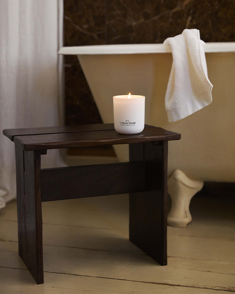 Marie-Stella-Maris No.13 L'Heure Santal Scented Candle burning on a dark wooden stool beside a freestanding bathtub. The warm bathroom setting creates a calming atmosphere inspired by the candle's signature fragrance with notes of sandalwood, cardamom and tonka.