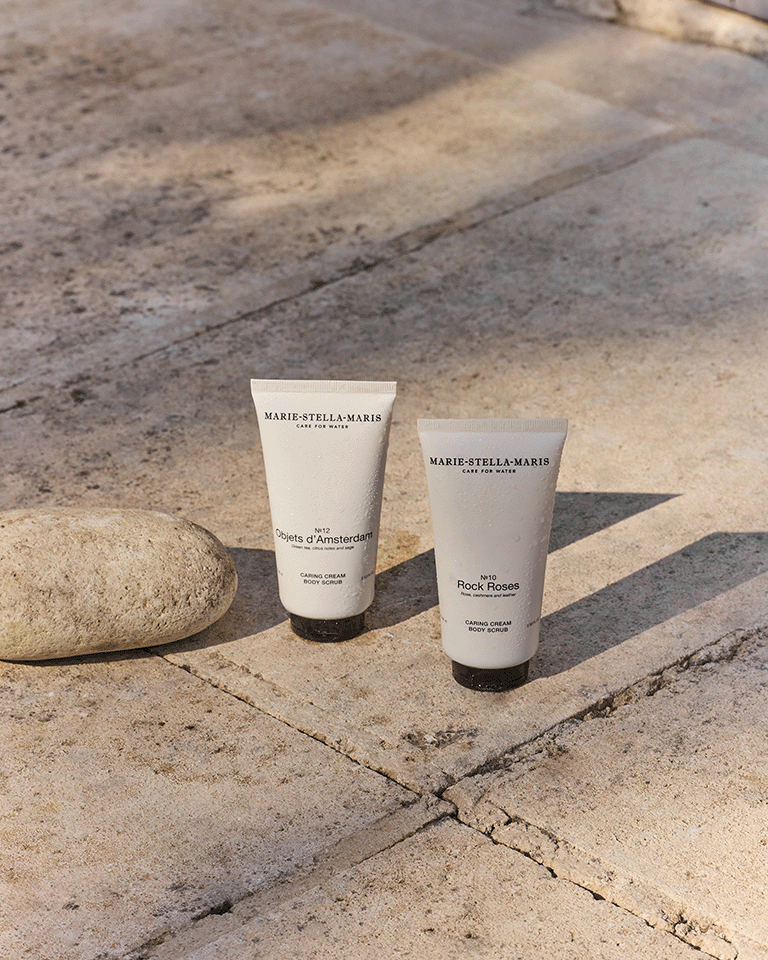 Marie-Stella-Maris Caring Cream Body Scrubs in Objets d’Amsterdam and Rock Roses fragrances displayed on a sunlit stone terrace beside a smooth natural rock. The minimalist white tubes cast long shadows, highlighting the products’ clean and premium design.