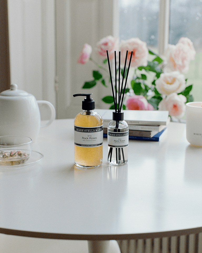 Marie-Stella-Maris No.10 Rock Roses products, including an exfoliating hand soap and fragrance sticks, styled on a table with tea and soft pink roses in the background, creating a serene setting for the Mother’s Day campaign.