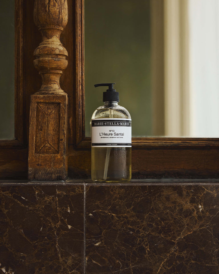 Marie-Stella-Maris No.13 L'Heure Santal Hand Soap (500 ml) displayed on a dark marble countertop beside a wooden-framed mirror. The elegant bathroom setting highlights the hand soap's minimalist design and its signature fragrance with notes of sandalwood, cardamom and tonka.