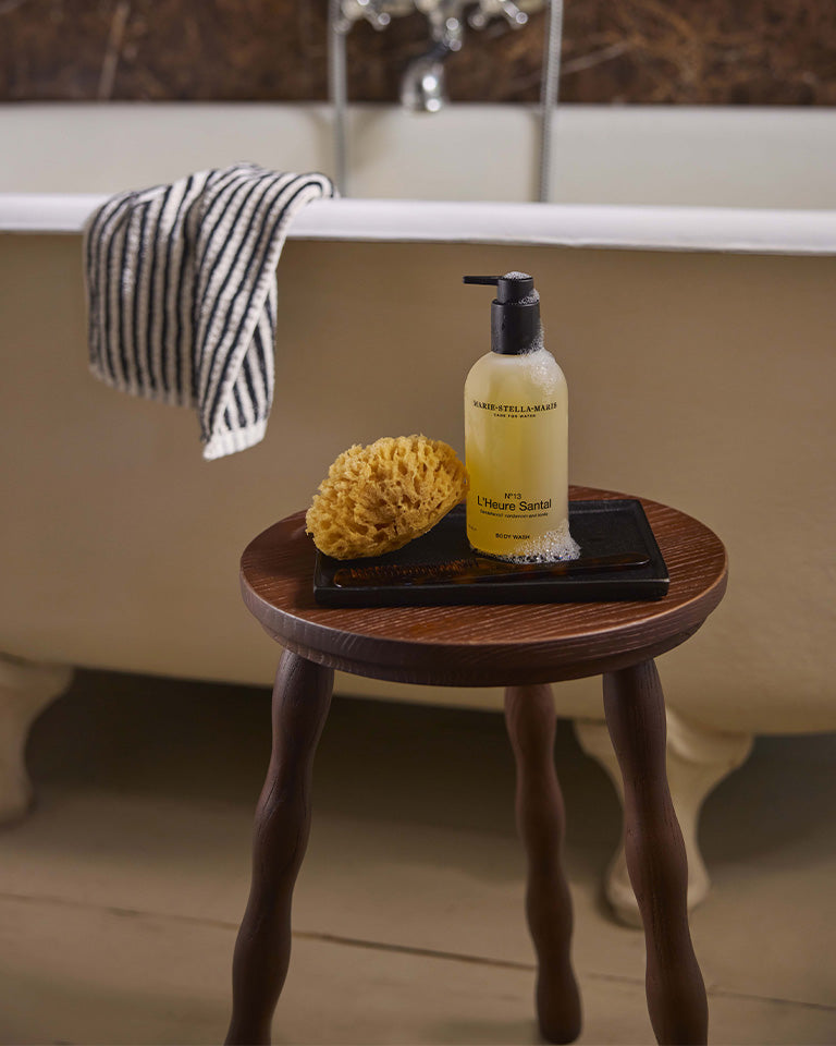 Marie-Stella-Maris No.13 L'Heure Santal Body Wash (300 ml) displayed on a dark tray with a natural sea sponge atop a wooden stool beside a freestanding bathtub. The warm bathroom setting highlights the body wash's signature fragrance with notes of sandalwood, cardamom and tonka.