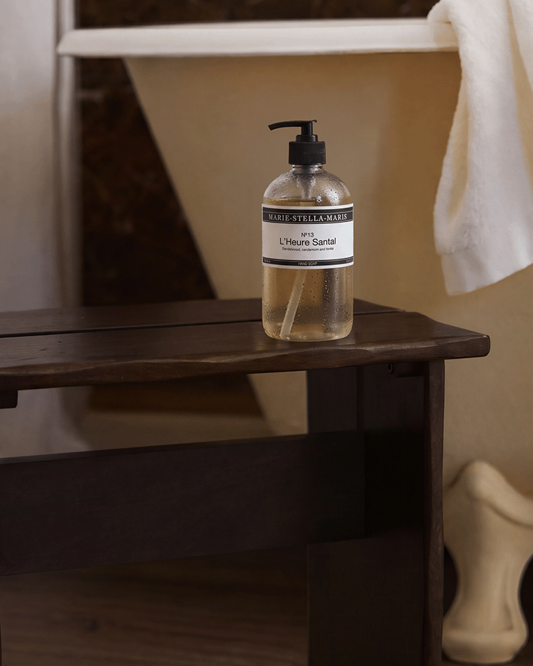 Marie-Stella-Maris No.13 L'Heure Santal Hand Soap (250 ml) displayed on a dark wooden bench beside a freestanding bathtub. Water droplets on the clear pump bottle and the warm bathroom setting evoke a fresh, calming hand care experience inspired by notes of sandalwood, cardamom and tonka.