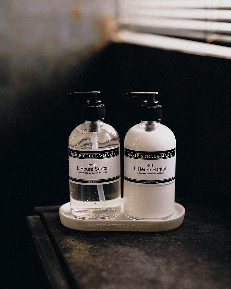 Marie-Stella-Maris No.13 L'Heure Santal Hand Soap and Hand Lotion displayed together on a white tray in a warm bathroom setting. The clear hand soap and white hand lotion bottles feature the signature fragrance with notes of sandalwood, cardamom and tonka, creating an elegant hand care duo.