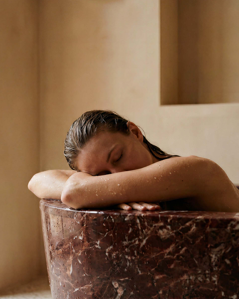 A person relaxes in a dark marble bath with their eyes closed, resting their arms on the edge of the tub. The warm, minimalist bathroom setting evokes a calm, spa-like atmosphere inspired by the comforting character of Marie-Stella-Maris No.13 L'Heure Santal.