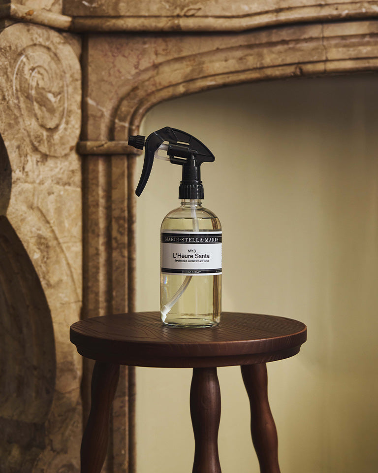Marie-Stella-Maris No.13 L'Heure Santal Room Spray (500 ml) displayed on a dark wooden side table in front of an elegant stone fireplace. The larger trigger spray bottle complements the warm interior, reflecting the fragrance's refined notes of sandalwood, cardamom and tonka.