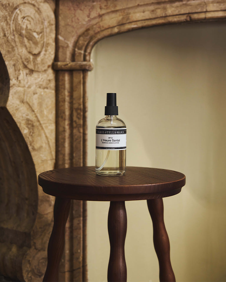 Marie-Stella-Maris No.13 L'Heure Santal Room Spray (100 ml) displayed on a dark wooden side table in a warm, elegant interior with a stone fireplace. The minimalist spray bottle complements the refined atmosphere inspired by the fragrance's notes of sandalwood, cardamom and tonka.