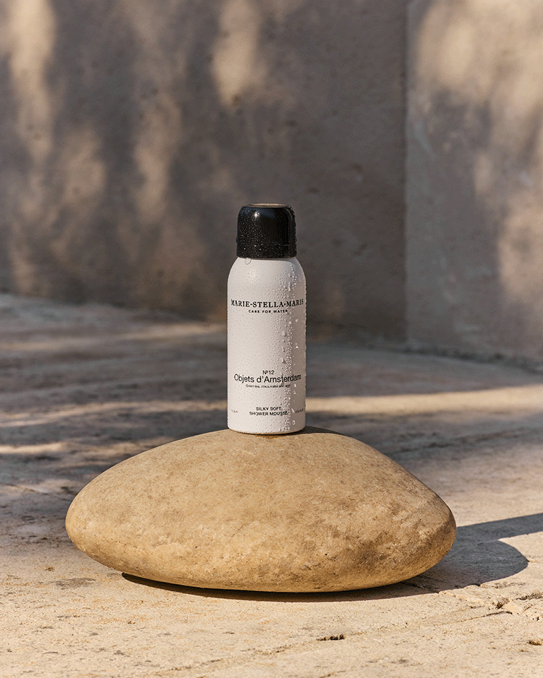 Marie-Stella-Maris No.12 Objets d’Amsterdam 75 ml travel-size Silky Soft Shower Mousse, placed on a smooth stone in a sunlit outdoor setting with soft shadows and water droplets on the bottle.