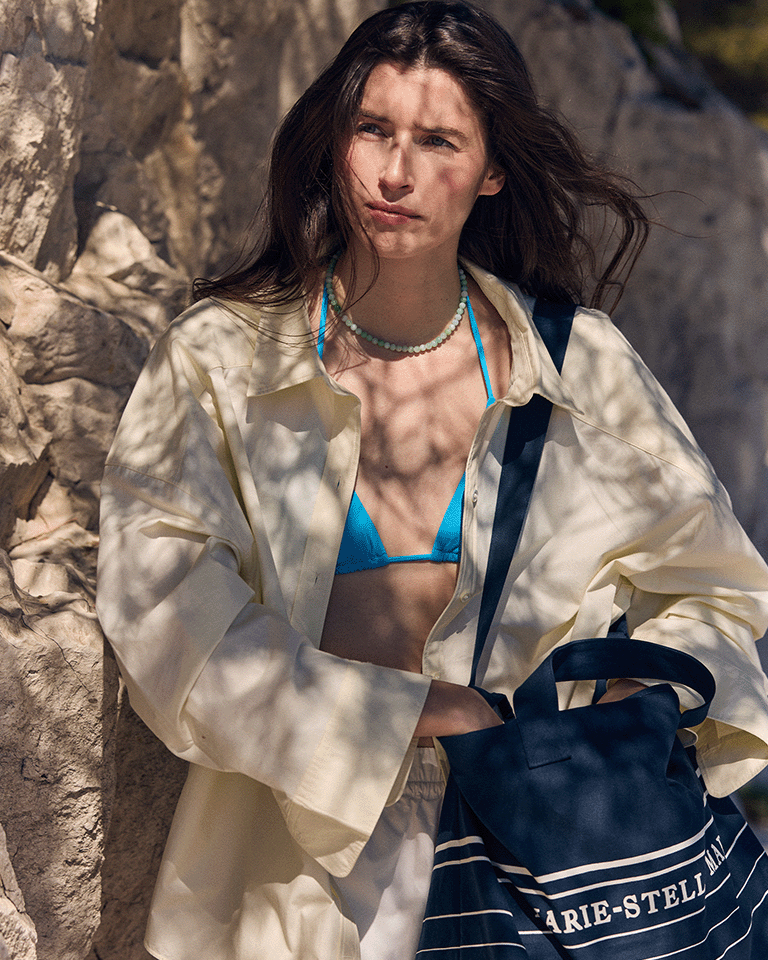Person wearing a blue bikini top and light shirt, holding a Marie-Stella-Maris limited edition summer 2026 navy blue tote bag with white stripe detailing, standing against a sunlit rocky backdrop with soft shadows.