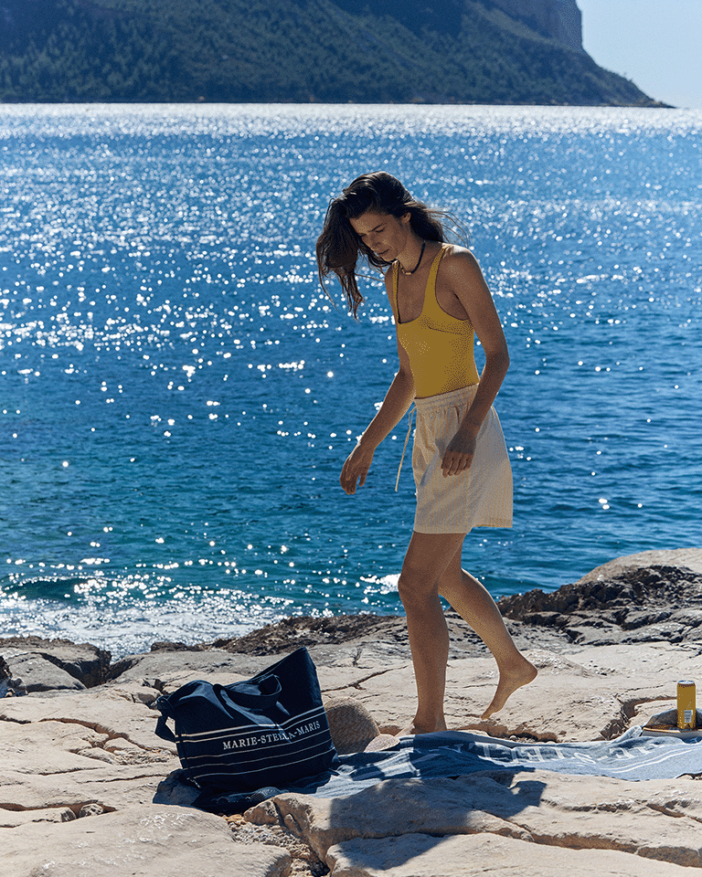 Person walking along a rocky beach wearing a yellow swimsuit and light shorts, next to a Marie-Stella-Maris limited edition summer 2026 navy blue tote bag and towel, with sparkling blue sea and coastline in the background.
