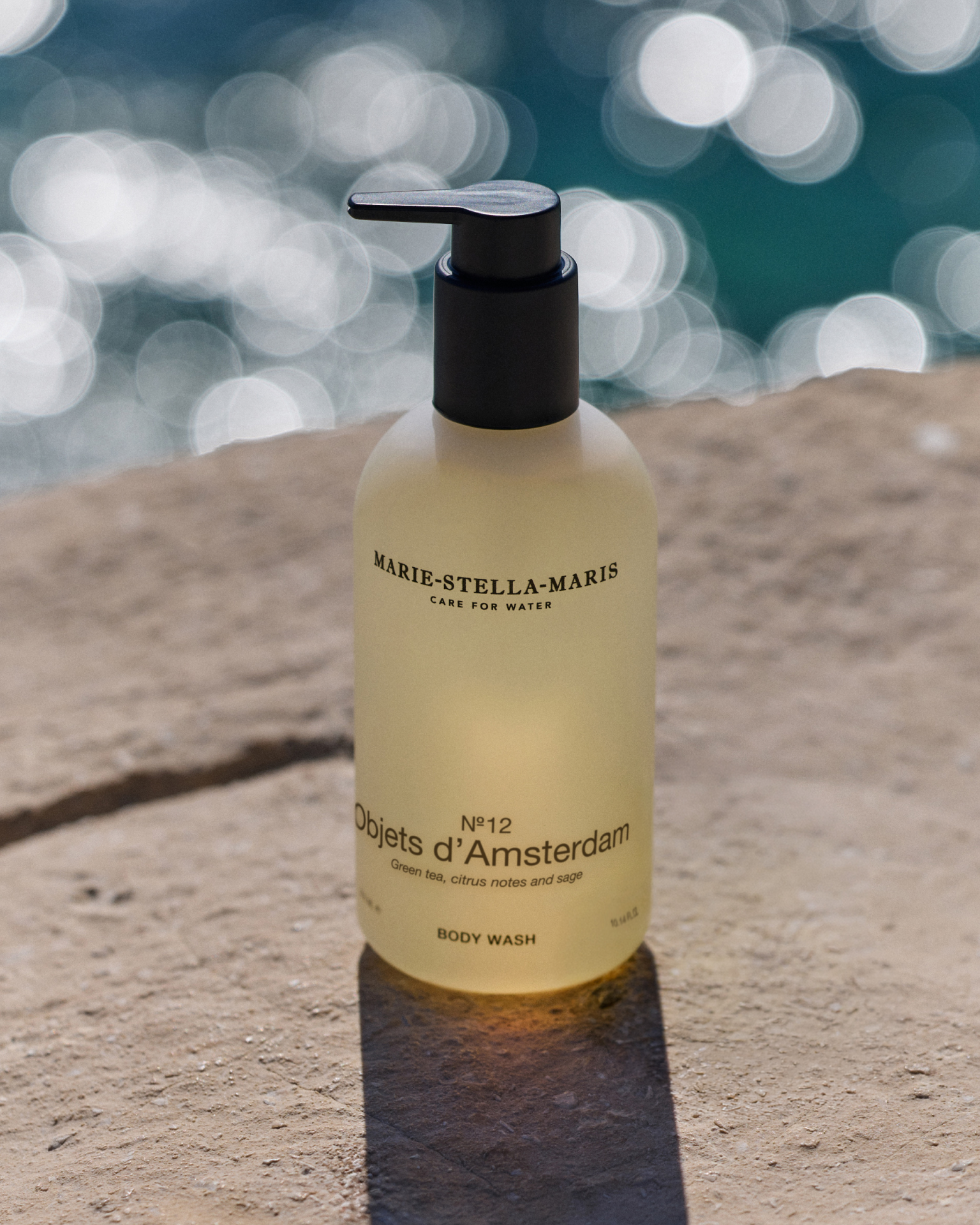 Marie-Stella-Maris No.12 Objets d’Amsterdam body wash 300 ml, featured as a May gift with purchase, displayed on a sunlit stone surface by the sea with sparkling water in the background.