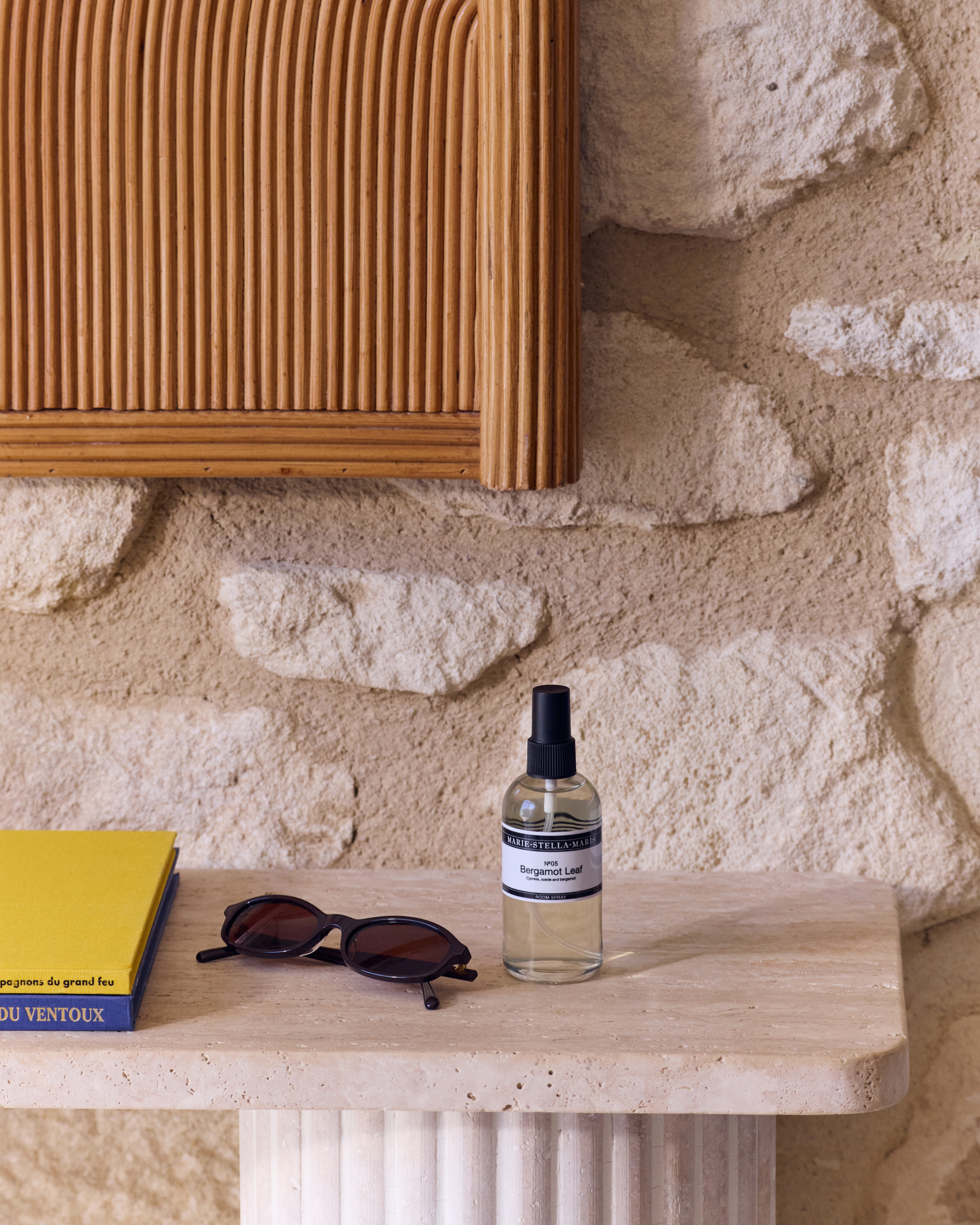 Marie-Stella-Maris No.05 Bergamot Leaf room spray styled on a stone table with sunglasses and books, set against a textured wall with a wooden mirror, creating a warm, summery interior scene.