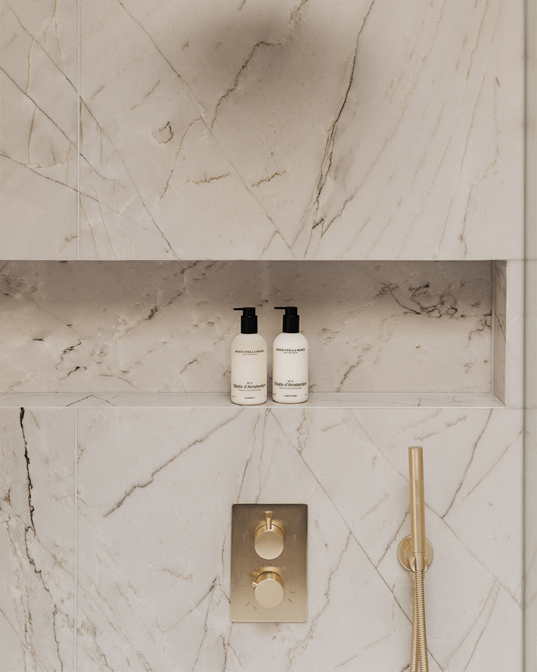 Two white Objets d'Amsterdam 300 ml conditioner bottles with black pumps on a marble shower shelf with gold fixtures