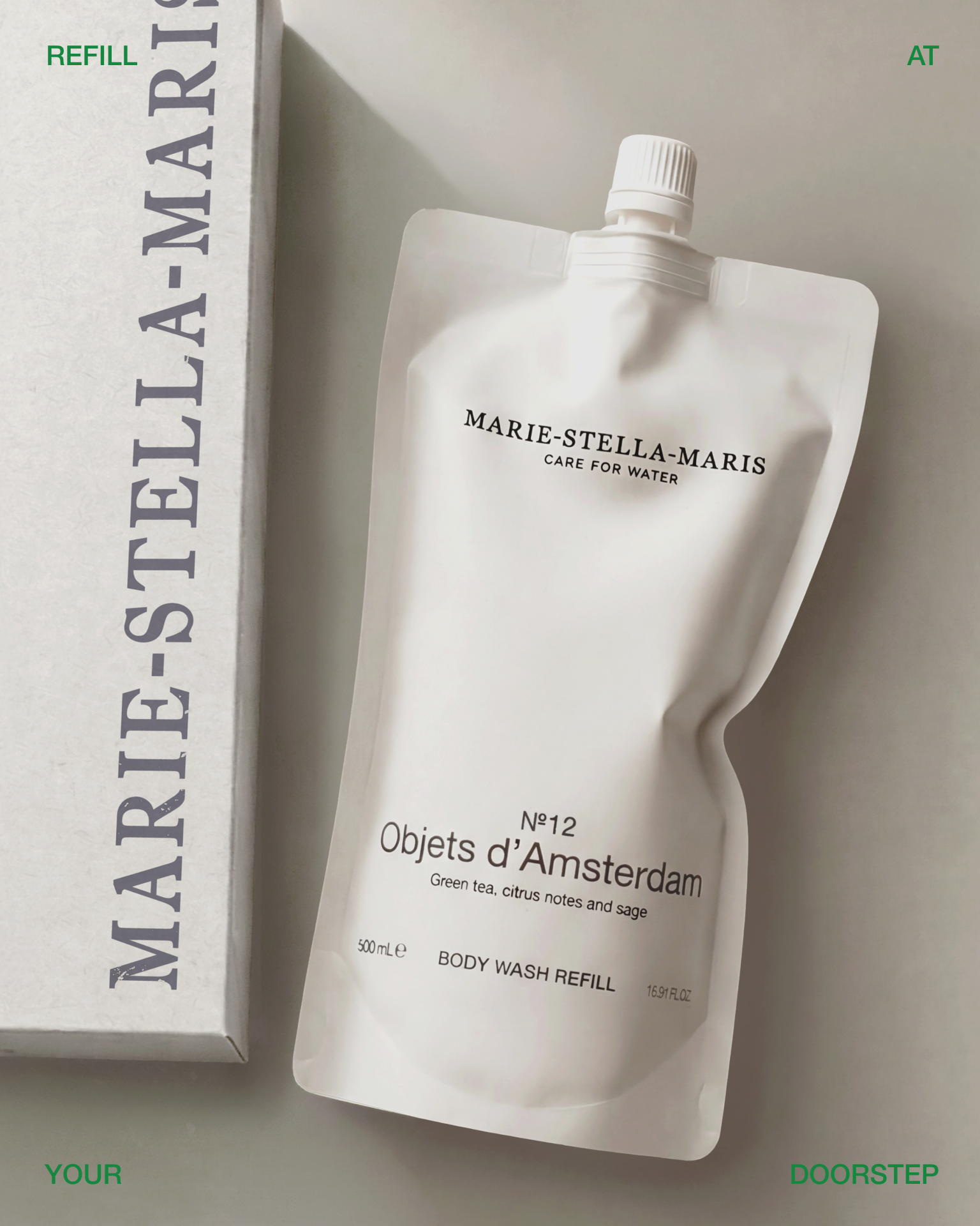 Marie-Stella-Maris No.12 Objets d’Amsterdam body wash refill pouch displayed beside a Marie-Stella-Maris shipping box on a soft neutral background. The image promotes the Marie-Stella-Maris refill subscription service, featuring a refill pouch delivered directly to your home for convenient, sustainable replenishment. Green text around the image reads “REFILL,” “AT,” “YOUR,” and “DOORSTEP.”