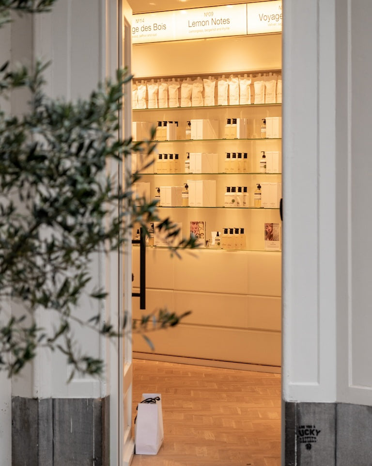 Looking through the doorway of the Marie-Stella-Maris store in Antwerp, Belgium—olive tree at the entrance, back-lit shelves of fragrances and refills, and a white shopping bag on the wooden floor.”