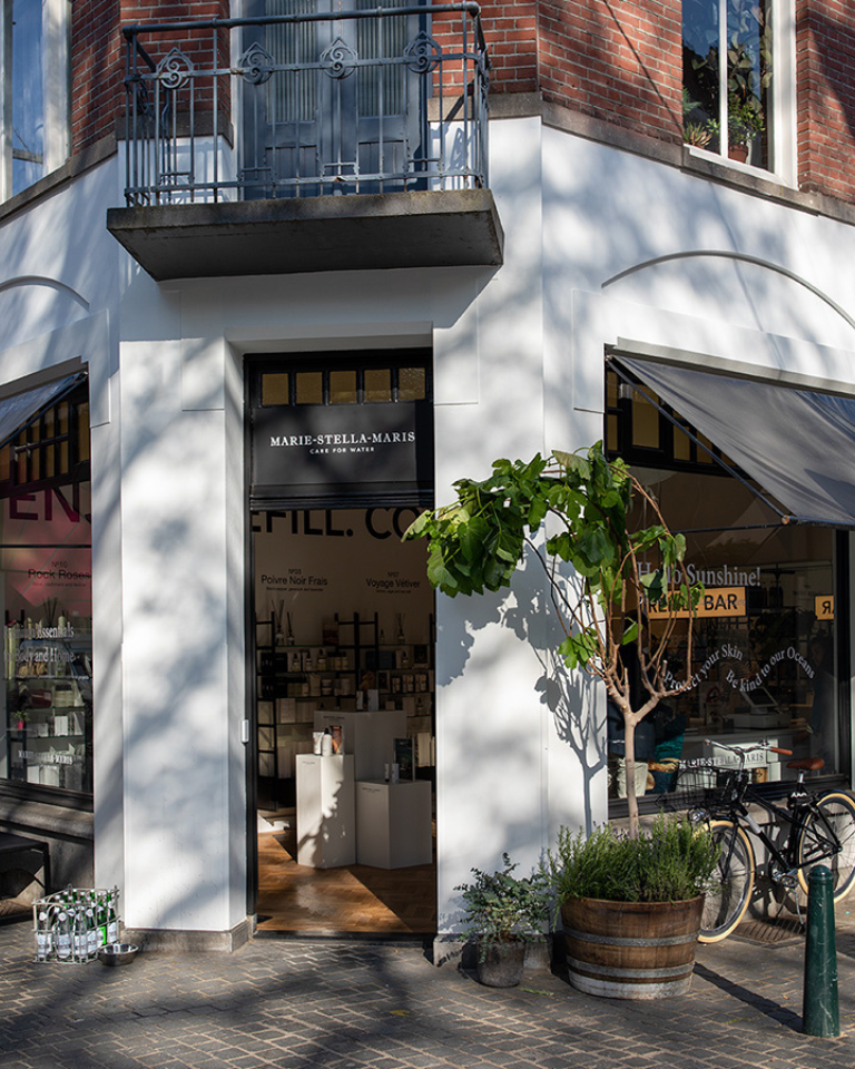 Corner storefront of the Marie-Stella-Maris boutique in Breda—white facade beneath an iron balcony, window awnings, planters and a bicycle outside, and ‘Refill Bar’ signage visible in the window.