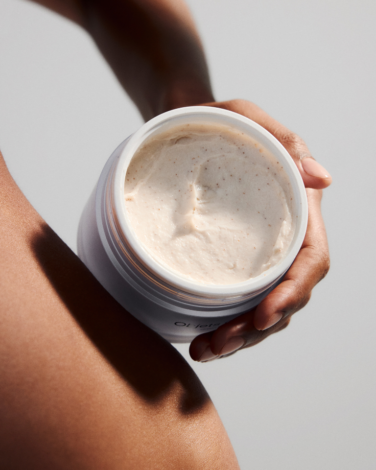 Open jar of Caring Cream Body Scrub with creamy texture held near smooth skin for gentle exfoliation and hydration