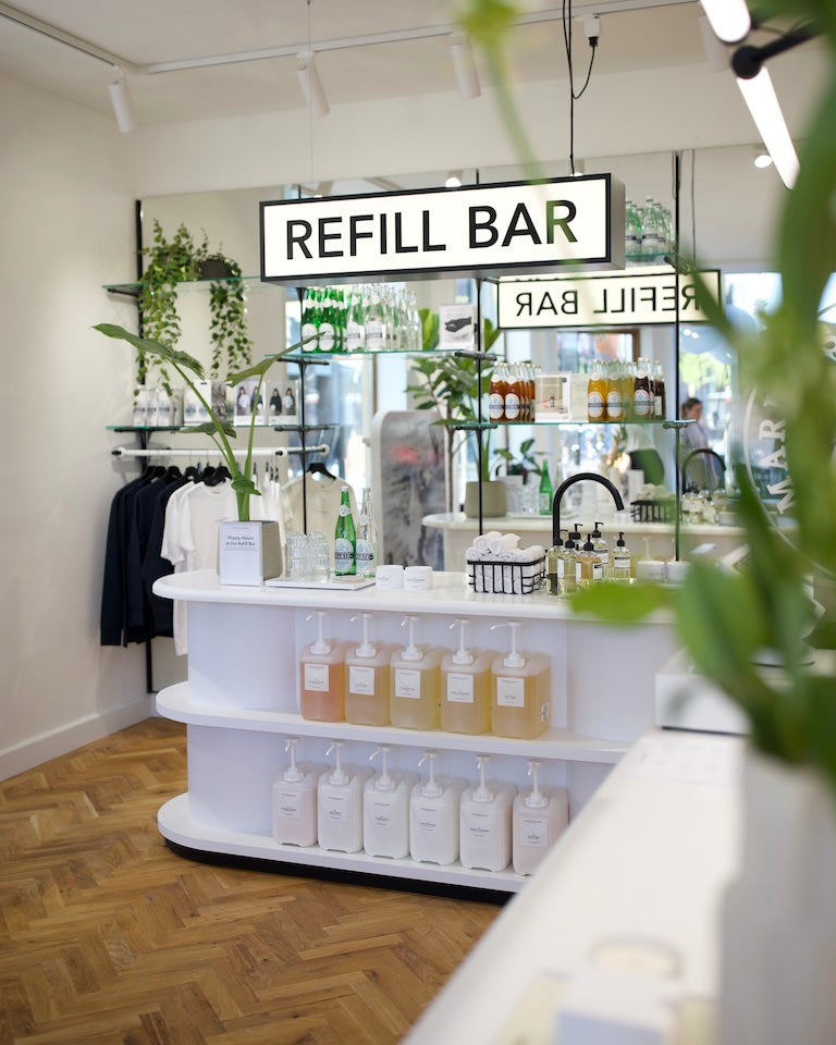 Inside the Marie-Stella-Maris store in Den Bosch (’s-Hertogenbosch): illuminated ‘Refill Bar’ sign over a curved white counter holding refill jerrycans and amber bottles, with mirrors, greenery, and herringbone wood flooring.