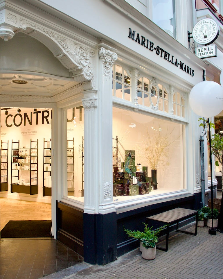 Corner facade of the Marie-Stella-Maris store in Den Haag (The Hague): ornate white trim over a dark base, large display window with product stacks, bench and planters outside, and a ‘Refill Station’ sign; view into the bright interior.