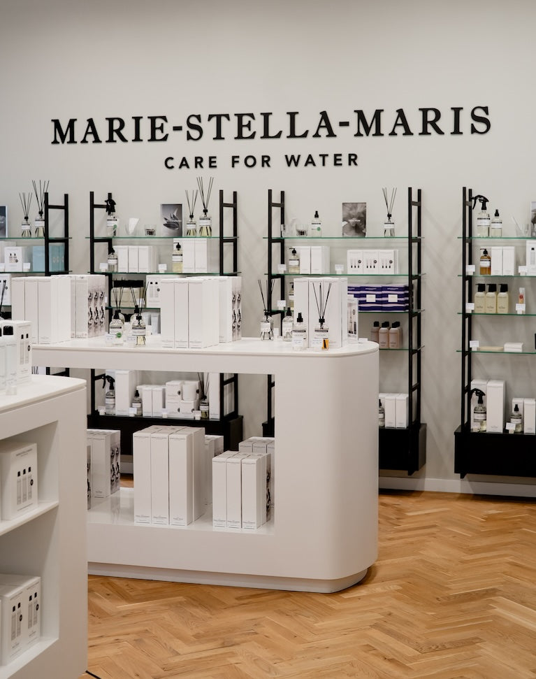 Inside the Marie-Stella-Maris boutique at Designer Outlet Roermond: large ‘MARIE-STELLA-MARIS – Care for Water’ wall sign above black-framed glass shelves of home fragrance and body care, with a curved white display island on herringbone flooring.