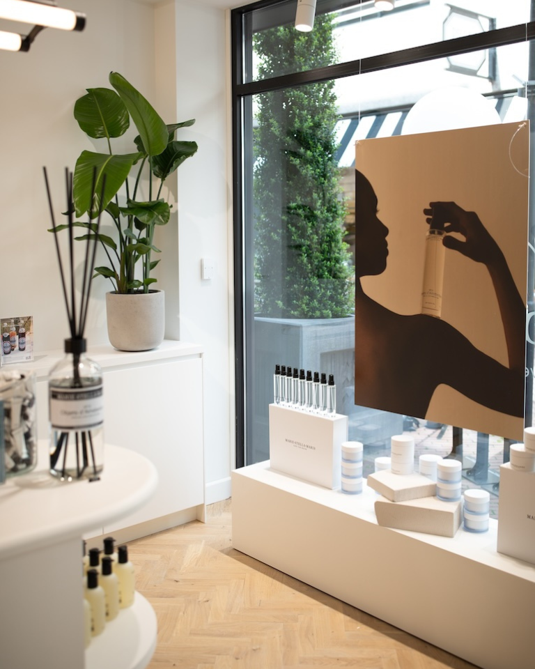 Inside the Marie-Stella-Maris store in Eindhoven: bright corner by a floor-to-ceiling window with a campaign poster (silhouette holding a bottle), white display plinths with products, reed diffuser, and a large potted plant on herringbone wood flooring.