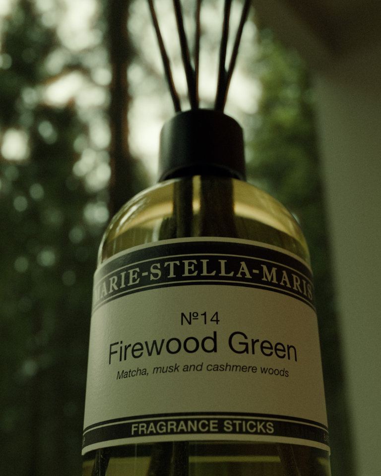 Close-up of Marie-Stella-Maris No. 14 Firewood Green fragrance sticks against a softly blurred woodland backdrop, evoking its notes of matcha, musk and cashmere woods.