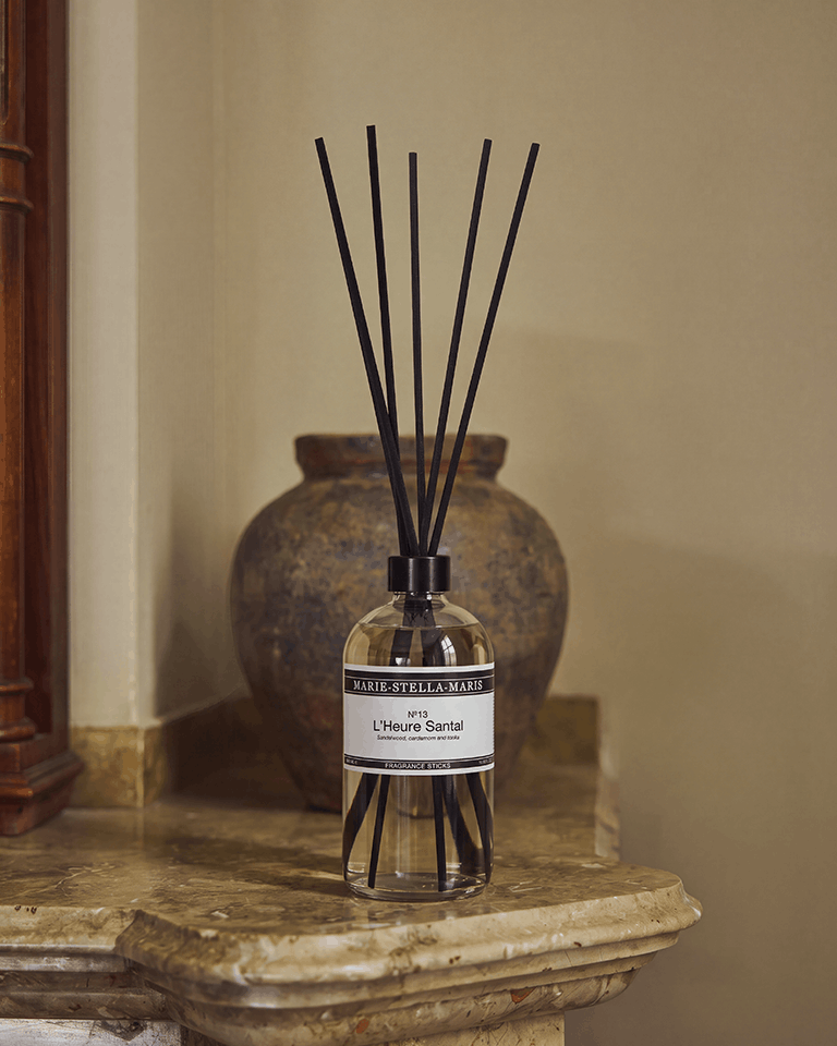 Marie-Stella-Maris No.13 L'Heure Santal Fragrance Sticks (500 ml) displayed on a marble mantelpiece beside a rustic ceramic vase. The clear glass diffuser with black fragrance reeds is showcased in a warm, elegant interior, reflecting the fragrance's refined notes of sandalwood, cardamom and tonka.