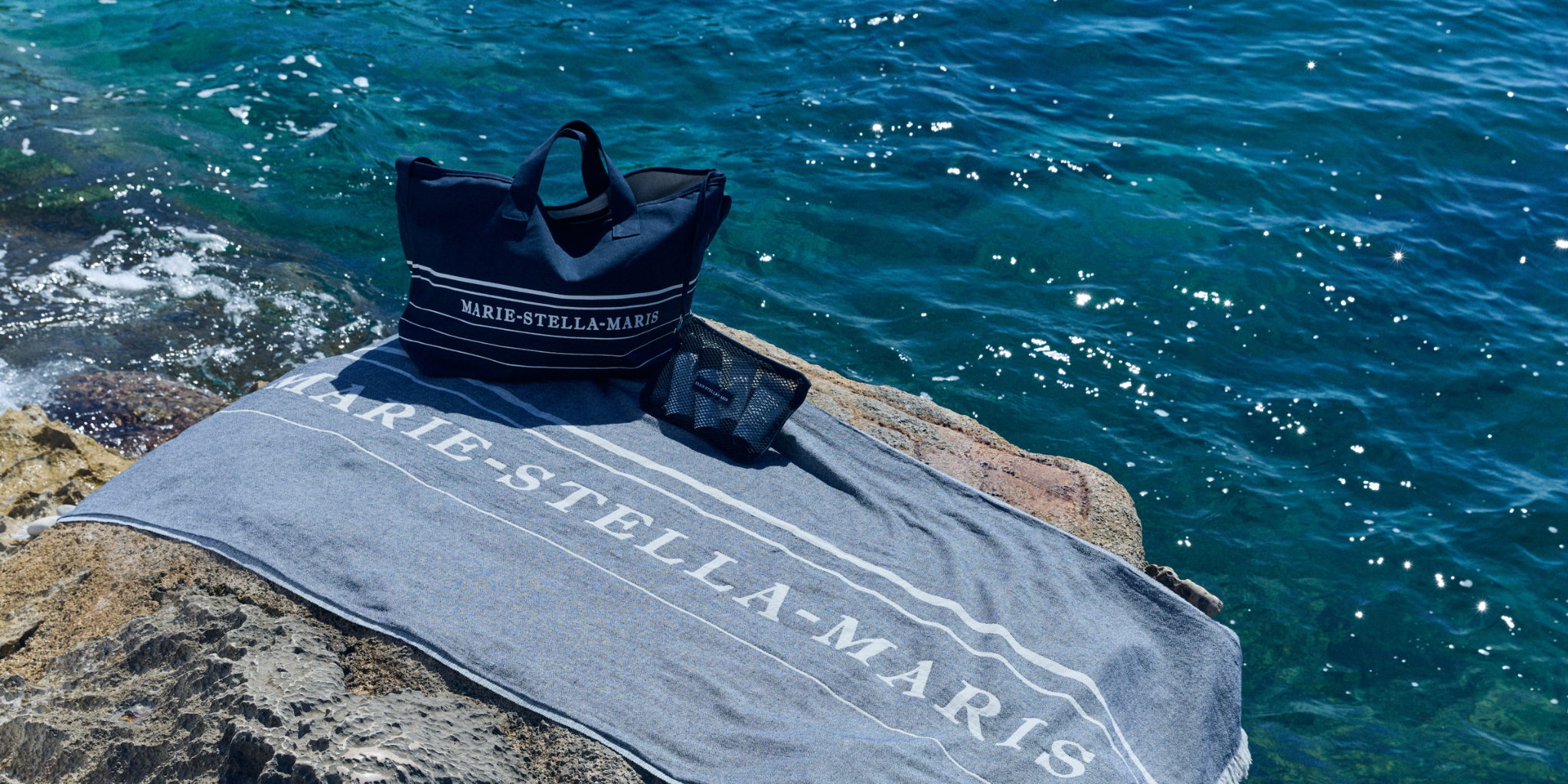 Marie-Stella-Maris summer essentials displayed on a rocky shoreline, featuring the limited edition gift with purchase collection: a navy blue summer tote bag, matching towel, and black mesh pouch, set against sparkling blue sea water.