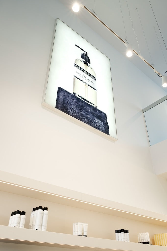 Interior of the Marie-Stella-Maris boutique in Bruges, featuring minimalist white shelving with products and a large illuminated fragrance display on the wall.
