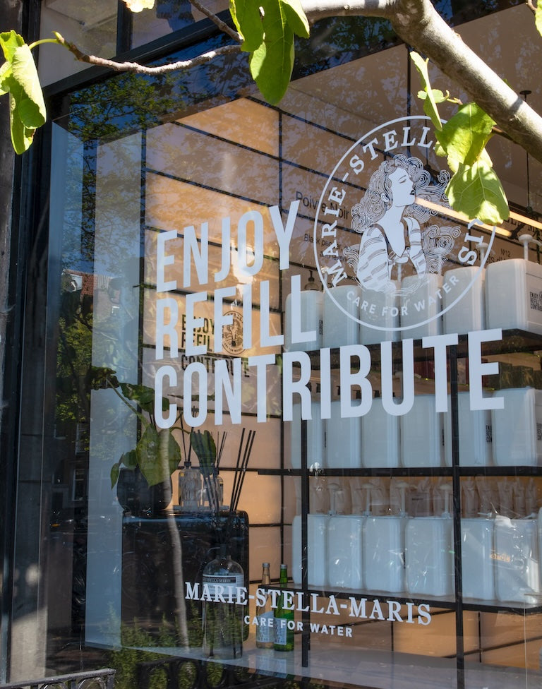 Close-up of the Marie-Stella-Maris Amsterdam Centrum storefront window showing the logo and ‘Enjoy Refill Contribute’ lettering, with refill canisters and bottles on shelves inside.