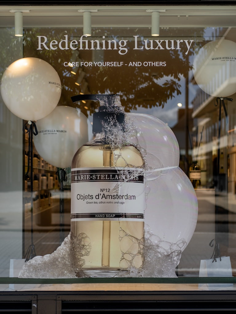 Marie-Stella-Maris storefront in Knokke, Belgium: large ‘Objets d’Amsterdam’ hand-soap bottle installation with soap bubbles and balloons; ‘Redefining Luxury’ tagline visible in the background.